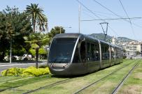 silver train running on train lines embedded in grass with green space and trees in the background 