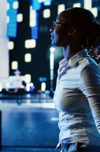 微笑的非裔美国妇女在夜晚漫步在城市的林荫大道上, 指着摩天大楼上有趣的广告牌. 在灯火照亮的街道上漫步的女商人