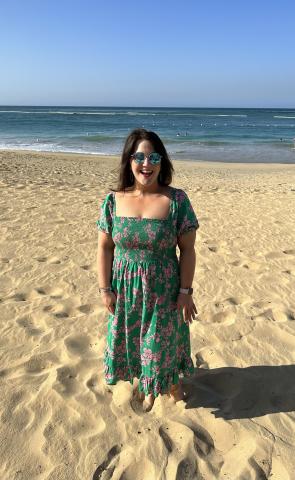一名黑发女子戴着墨镜，身穿绿色连衣裙，在沙滩上以海洋为背景摆姿势