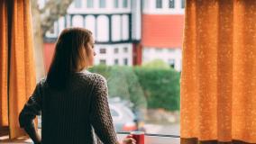 Stock image of woman in a beige sweater looking out a window draped with gold curtains