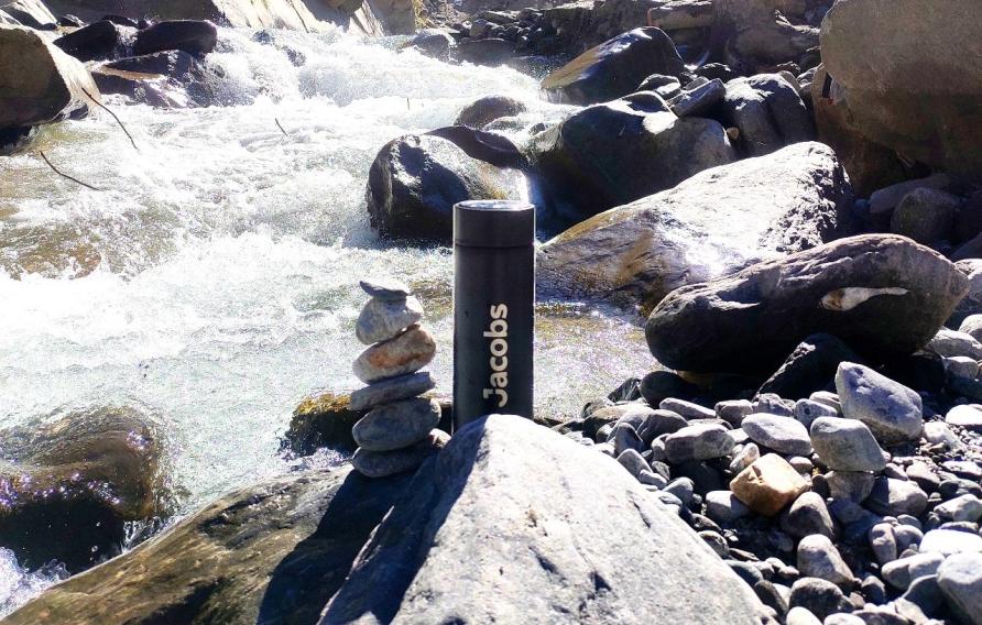 Black Jacobs water bottle sitting next to piled rocks in a river with greenery and blue sky background