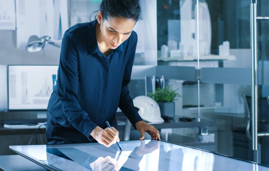 Talented Young Female Architectural Designer Draws Building Concept on a Graphics Tablet Display. Clean Minimalistic Office, Concrete Walls Covered by Blueprints.