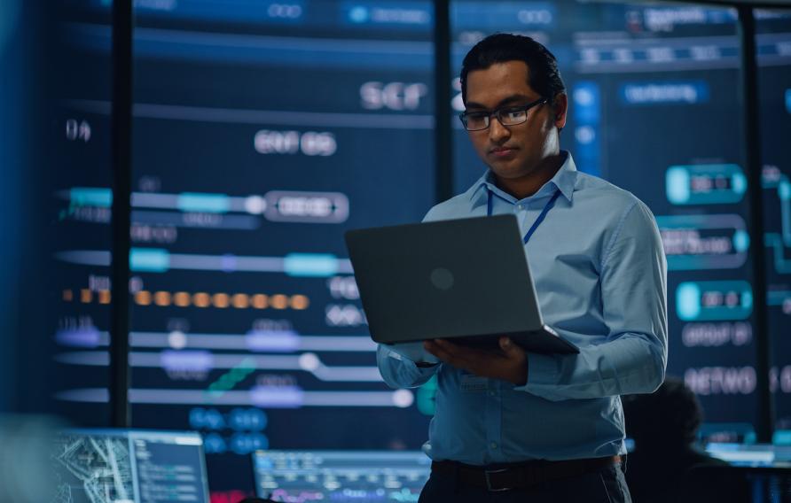 Young Multiethnic Male Government Employee Uses Laptop Computer in System Control Monitoring Center. In the Background His Coworkers at Their Workspaces with Many Displays Showing Technical Data.