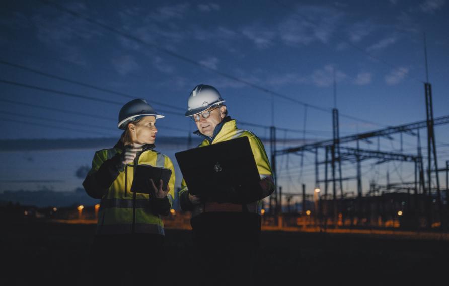 Electrical Engineers Discussing Over Laptop And Digital Tablet While Standing At Dark Power Station During Night stock photo