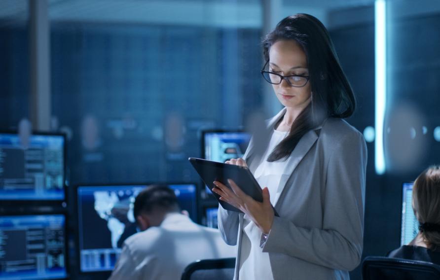 Young Female Engineer Uses Tablet in System Control Center. In the Background Her Coworkers are at Their Workspaces with many Displays Showing Valuable Data.