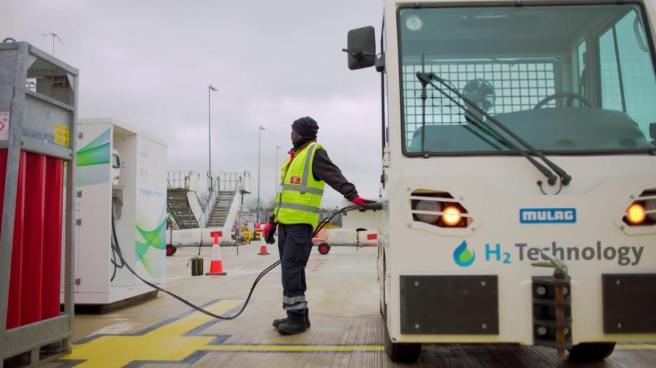 Man in hi viz yellow waistcoat refueling a luggage tug at an airport.