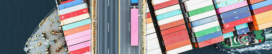 Description Aerial view of a container ship passing beneath a suspension bridge. Semi truck with pink cargo container crosses above.